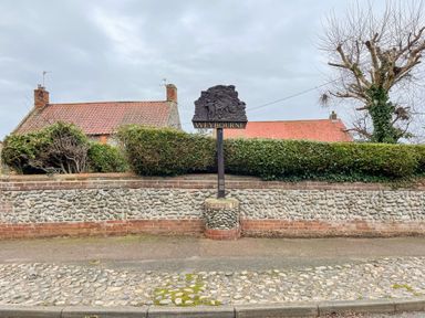 A stone and brick wall with bushes and a tree behind it and a sign that says Weybourne in front of houses with tiled roofs at Timbers in Weybourne