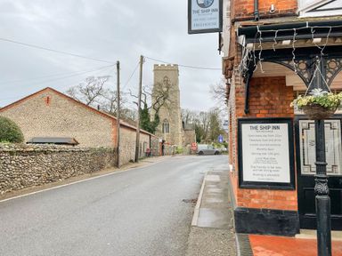 Street view with a pub corner sign outside The Ship Inn and a church tower in the background in Weybourne