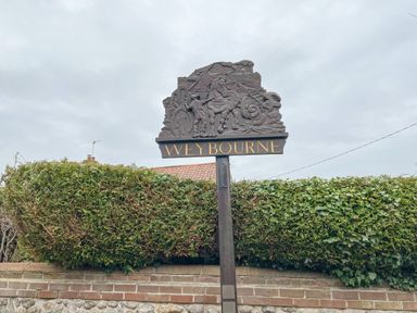 A wooden village sign with relief carvings and the word Weybourne surrounded by greenery and a brick wall