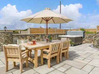 An outdoor patio with a wooden dining table and chairs under a large umbrella and a hot tub against a stone wall at Refail Fach Near Cemaes Bay