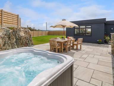 An outdoor patio with a hot tub wooden dining table with six chairs and an umbrella near a dark gray building at Refail Fach Near Cemaes Bay