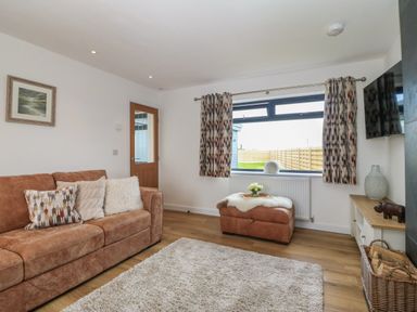 A living room with a brown sofa and patterned cushions a window with patterned curtains an ottoman with a tray and flowers a wooden door and a TV mounted on the wall at Refail Fach Near Cemaes Bay