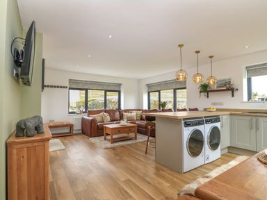 An open plan living room and kitchen with a brown leather corner sofa a wooden coffee table a TV on the wall and washing machines under the kitchen counter at Refail Fach Near Cemaes Bay