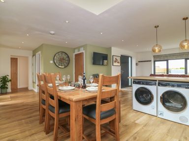 A dining area with a wooden table and six chairs set with plates and glasses next to a kitchen island with a washing machine and dryer at Refail Fach Near Cemaes Bay