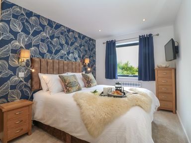 A bedroom with a bed with pillows and a sheepskin on it two wooden bedside tables blue curtains and a wall-mounted television at Refail Fach Near Cemaes Bay