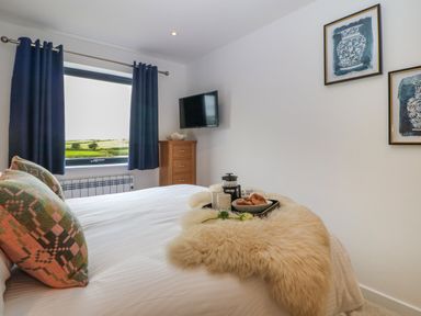 A bedroom with a white bed covered with a fluffy throw and food tray two framed pictures on the wall a wooden chest of drawers under a window with blue curtains and a wall mounted TV at Refail Fach Near Cemaes Bay