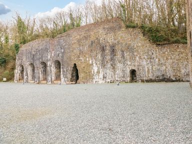 A stone wall with arched openings and a gravel ground with trees in the background at Dono's Lodge in Stepaside near Kilgetty Heritage Park
