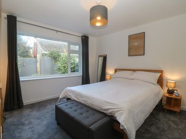 A bedroom with a double bed white bedding a gray ottoman at the foot of the bed two lamps wooden bedside table and a large window with curtains at Ty Nain in Llanfairpwllgwyngyll