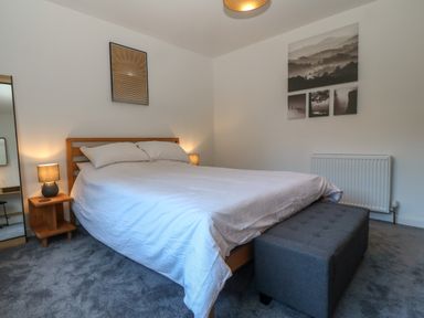 A bedroom with a double bed white bedding wooden headboard bedside table with lamp gray cushioned bench and framed pictures on the wall at Ty Nain in Llanfairpwllgwyngyll