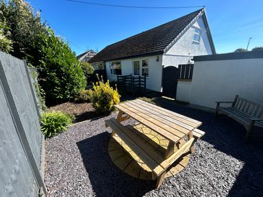 An outdoor area with a wooden picnic table and bench next to a white building with dark roof and a garden at Ty Nain in Llanfairpwllgwyngyll