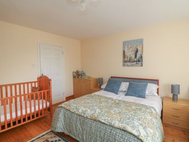 A bedroom with a double bed, two bedside tables with lamps, a crib with a teddy bear, a cabinet with books, and a cityscape painting on the wall at Bolger House in Ballintubbrid near Kilmuckridge County Wexford