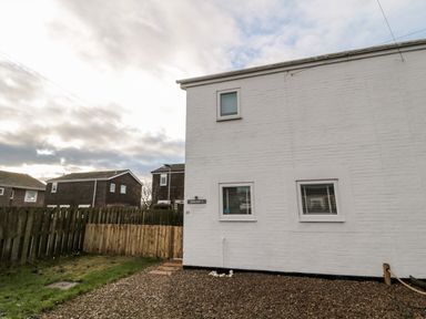 The exterior side of a white painted house with three windows and a gravel area next to a fenced grassy yard at 22 Longstone Park in Beadnell