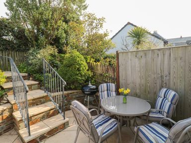 An outdoor patio with four cushioned chairs around a table with flowers and a barbecue grill next to stairs and plants at Bumbles in Padstow