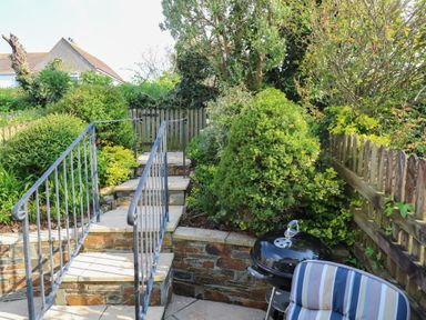 A stone patio with a black barbecue grill a striped chair and steps with a metal railing leading up to a wooden fence surrounded by shrubs and trees at Bumbles in Padstow