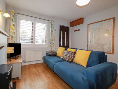A living room with a blue sofa with yellow cushions a wooden floor a small TV on a table a window with patterned curtains and a wall map at Bumbles in Padstow