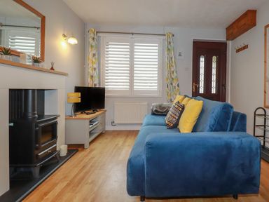 A living room with a blue sofa yellow pillows a wood stove a television on a stand and a window with patterned curtains at Bumbles in Padstow