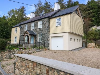 A stone and siding house with a garage and gravel driveway surrounded by trees at Belle View Llaniestyn near Abersoch