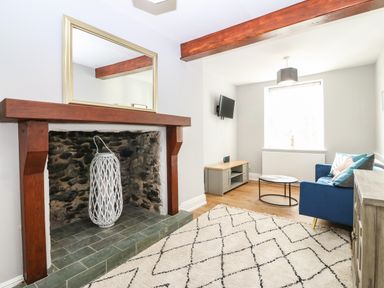 A living room with a stone fireplace wooden beams a blue sofa a round coffee table a TV mounted on the wall and a patterned rug at Belle View in Llaniestyn near Abersoch