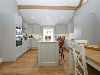 A kitchen with light gray cabinets an island with brown bar stools and a wooden dining table at Belle View Llaniestyn near Abersoch