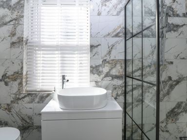 A bathroom with a white sink on a vanity unit a window with white blinds and a glass shower enclosure at Belle View in Llaniestyn near Abersoch