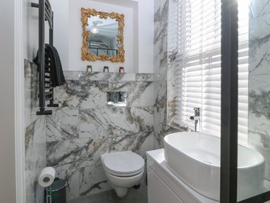 A bathroom with a wall-mounted toilet a white oval sink a black towel rack and a window with blinds at Belle View in Llaniestyn near Abersoch