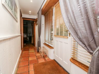 A hallway with terracotta floor tiles a white door with wooden blinds grey curtains a small wooden shoe rack and a framed picture on the wall at The Shippen in Gooseham near Kilkhampton