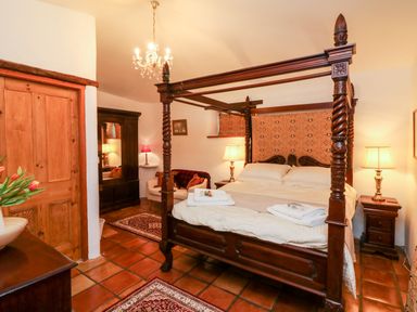 A bedroom with a four-poster bed two bedside tables with lamps a wooden wardrobe an armchair and a chandelier at The Shippen in Gooseham near Kilkhampton