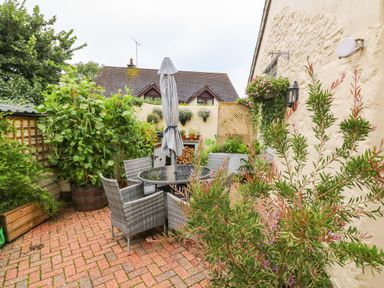An outdoor patio area with a glass table and four wicker chairs surrounded by various potted plants and a closed umbrella at The Shippen in Gooseham near Kilkhampton