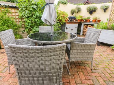 A patio with a round glass table and four wicker chairs surrounded by potted plants and a storage unit at The Shippen in Gooseham near Kilkhampton