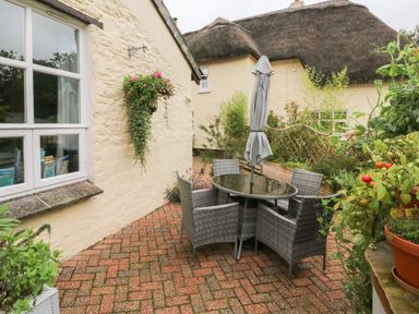 An outdoor patio area with a round glass table surrounded by four wicker chairs and a closed umbrella next to a stone wall with a hanging flower basket at The Shippen in Gooseham near Kilkhampton
