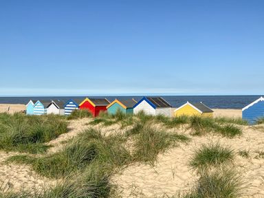 A sandy beach with grass and colorful beach huts near the sea at Sea Mouse Cottage in Southwold