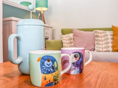 A wooden table with two colorful bird illustrated mugs and a light blue French press with a sofa and cushions in the background at Sea Mouse Cottage in Southwold