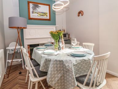 A dining table set with plates glasses and cutlery surrounded by white chairs near a fireplace with a framed picture above at Sea Mouse Cottage in Southwold