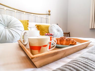 A breakfast tray with cups, a croissant, and jam on a bed with pillows at Sea Mouse Cottage in Southwold