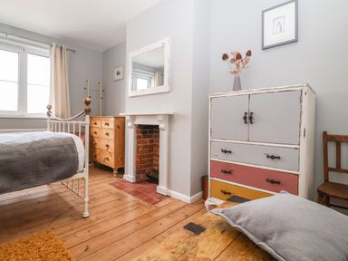 A bedroom with a metal bed frame wooden floors a wooden chest of drawers a small fireplace a multicolored cabinet a wooden chair and a window with curtains at Sea Mouse Cottage Southwold