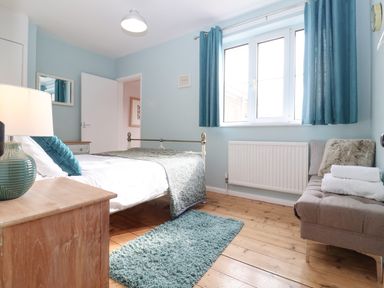A bedroom with a bed bedside table mirror window with blue curtains and a cushioned bench with towels at Sea Mouse Cottage Southwold