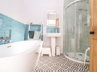 A bathroom with a white bathtub blue tiled walls a white sink with mirror a glass shower enclosure and patterned floor tiles at Sea Mouse Cottage Southwold