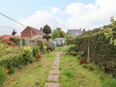 A garden pathway with grass and plants on both sides leading to a small shed and houses in the background at Sea Mouse Cottage in Southwold