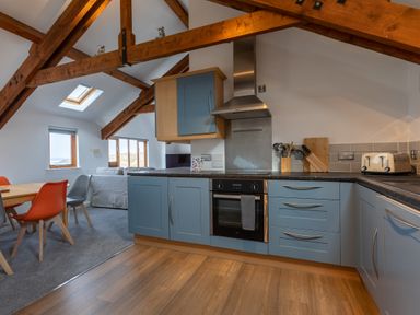 A kitchen with blue cabinets and wooden beams next to a dining area with chairs and a sofa at Sunset View in Widemouth Bay