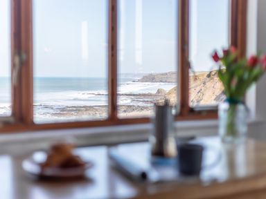A coastal view from a window with table in foreground holding a cup flowers and plate at Sunset View in Widemouth Bay