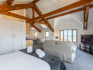 A room with a bed with towels a seating area a dining table wooden ceiling beams and large windows showing the sea at Sunset View in Widemouth Bay