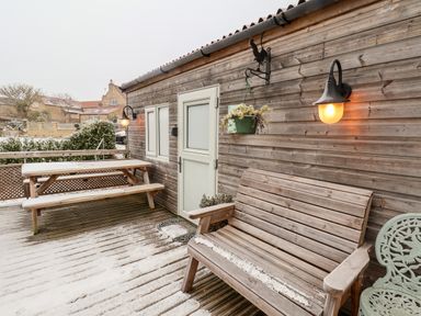 An outdoor wooden deck with a bench, picnic table, hanging plant, wall lights, and a white door at Grouse Lodge in Seamer, North Yorkshire