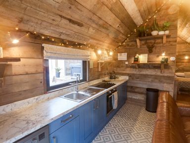 A kitchen with blue cabinets marble countertop and wooden walls with shelves holding plants and glasses at Foxes Den Seamer North Yorkshire