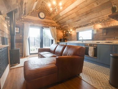 A living room with a brown leather sofa near a kitchen area with blue cabinets and a wood burning stove at Foxes Den in Seamer North Yorkshire