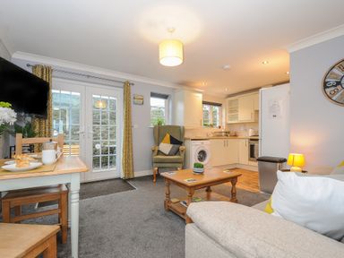 An open living room and kitchen with a sofa, armchair, coffee table, dining table set near French doors and kitchen cabinets at 1 Bethany Court in Bodmin