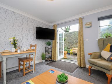 A living room with a small dining table set for breakfast a television mounted on the wall and French doors leading to an outdoor area at 1 Bethany Court in Bodmin