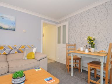 A living room with a beige sofa pillow and a wooden table with plants and coasters and a dining table set with flowers and breakfast at 1 Bethany Court in Bodmin