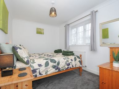 A bedroom with a wooden bed, floral bedding, a nightstand with a lamp, a window with curtains, and a dresser with a mirror and plant at 1 Bethany Court in Bodmin