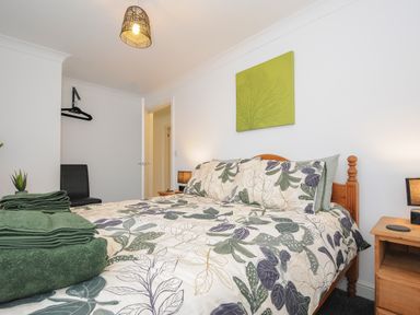 A bedroom with a double bed with floral bedding green towels on the bed wooden bedside tables with lamps a black chair and a green wall art at 1 Bethany Court in Bodmin