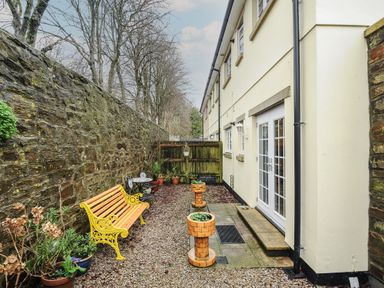 An outdoor patio area with a yellow bench small table and chairs potted plants and two brick planters at 1 Bethany Court in Bodmin